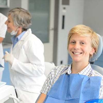 A young boy smiling in the dentist’s chair while the dentist and assistant review paperwork in the background