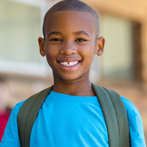 A little boy wearing a blue shirt and backpack smiling while at school