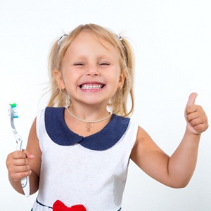 A little girl with blonde hair smiling and giving a thumbs-up while holding a toothbrush after receiving fluoride treatment for kids in McKinney