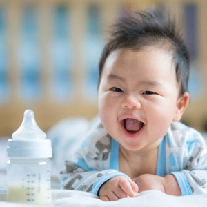 A baby lying on its stomach and smiling while a bottle of milk sits within arm’s reach