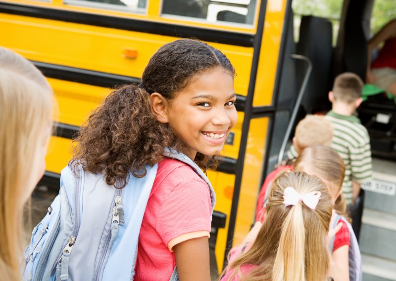 Girl smiling with teeth as she prepares to get on yellow school bus with other children